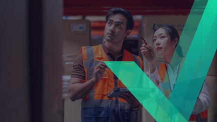 Two individuals wearing orange safety vests review information on a tablet in a warehouse setting, surrounded by stacked boxes.