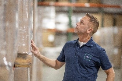Warehouse worker in blue polo shirt inspecting a wrapped pallet among shelves and boxes.